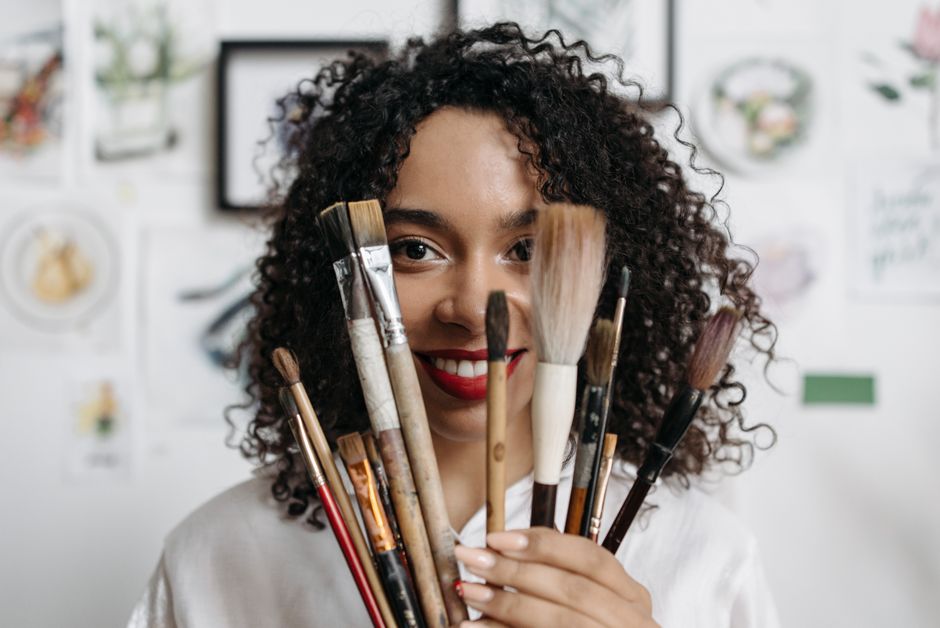 Artistic young woman with curly hair smiling while holding paintbrushes indoors.