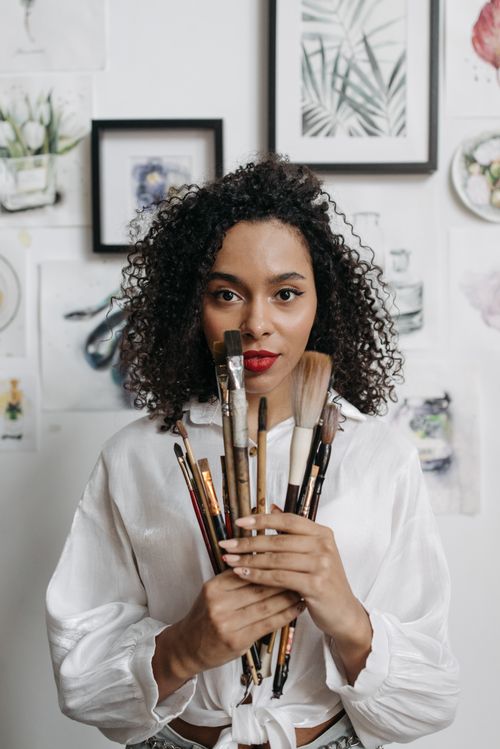 Free Portrait of a black female artist holding paintbrushes, surrounded by art. Stock Photo