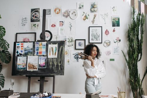 Free Artistic woman with brushes in studio surrounded by paintings and greenery. Inspiring atmosphere. Stock Photo