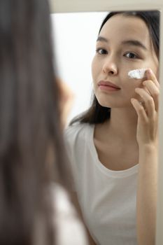 Young woman applies skincare product on face while looking at her reflection in the mirror.
