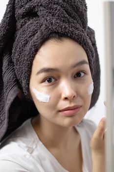 Young woman in towel wrap applying face cream, indoors, skincare routine.