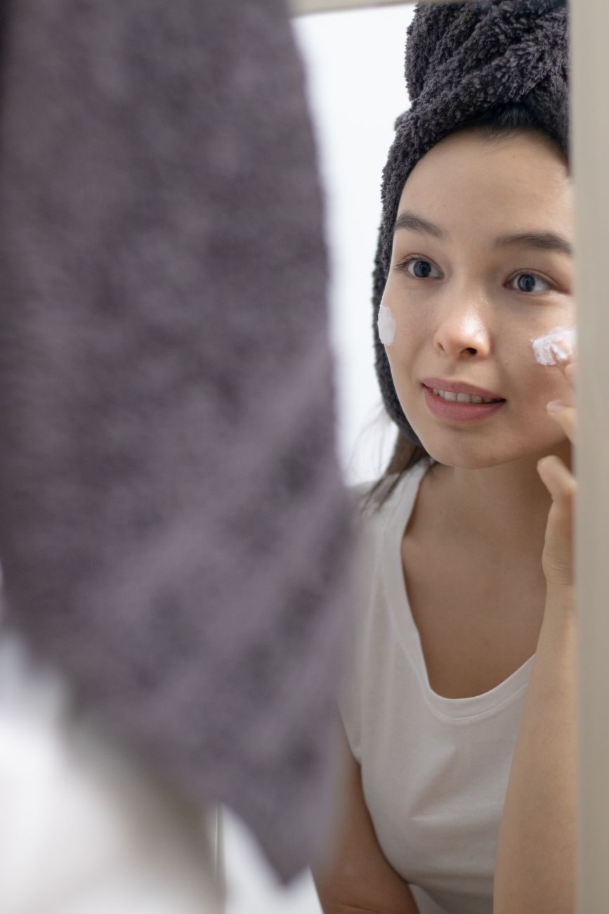 Woman with a towel on her head applying moisturizer in front of a mirror for skincare routine.
