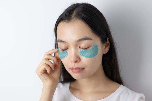 Free Serene Asian woman with closed eyes applying blue under-eye patches for skincare routine. Stock Photo