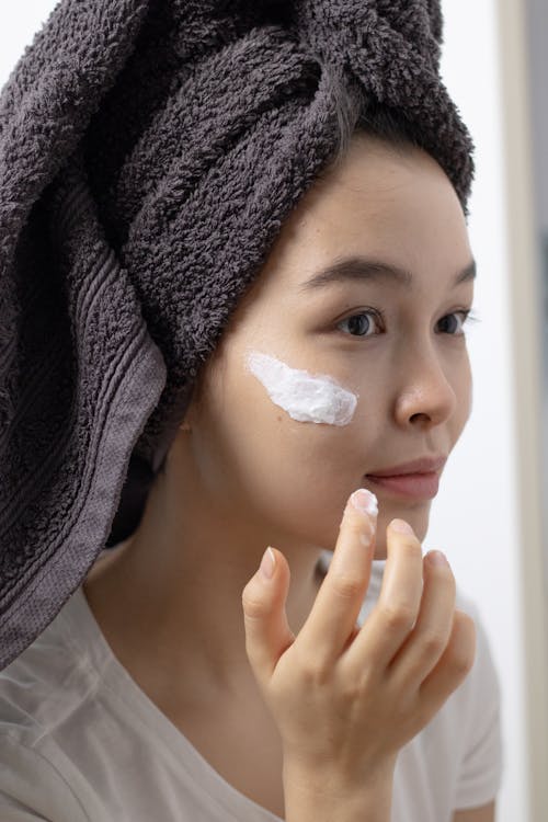 
A Woman with a Towel on Her Head Applying a Skin Care Product on Her Face 