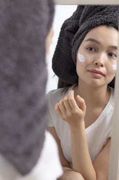 Woman in towel applying face cream, looking at her reflection. Skincare routine in the bathroom.