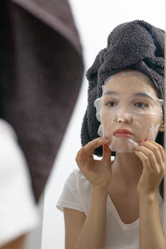 Young woman applying a hydrating sheet mask in front of a bathroom mirror.