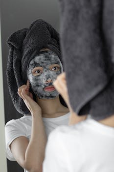 Young woman with towel-wrapped hair applying face mask, reflected in a mirror.