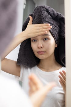 Young woman in front of mirror practicing skincare routine with head towel indoors.