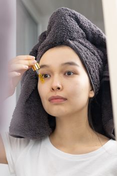 An Asian woman applies skincare serum with a dropper in front of a mirror.