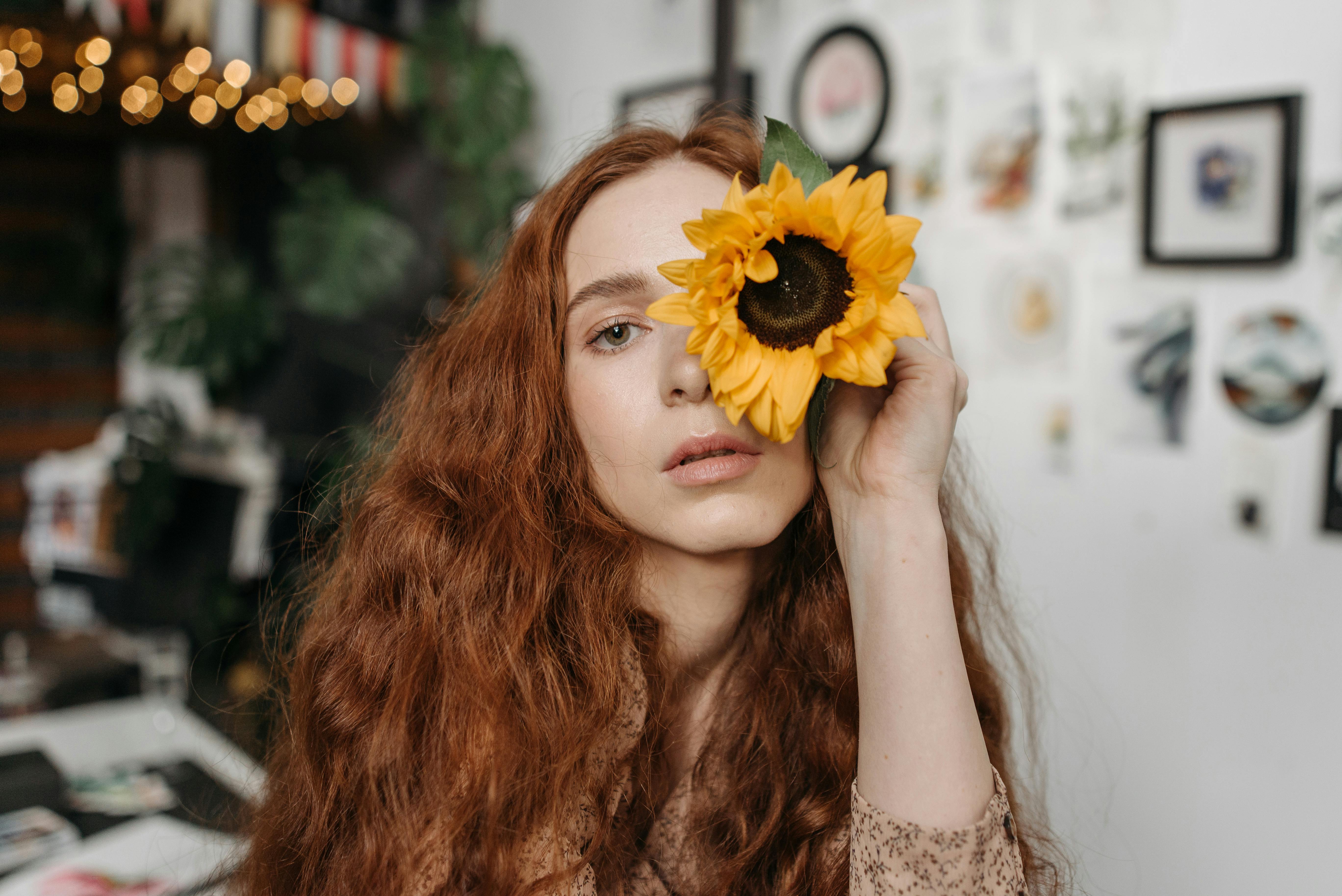 Portrait of a redheaded woman with a sunflower covering one eye, set indoors with artistic decor.