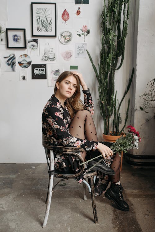 A Woman in Black Floral Dress Sitting on the Chair while Holding Flowers