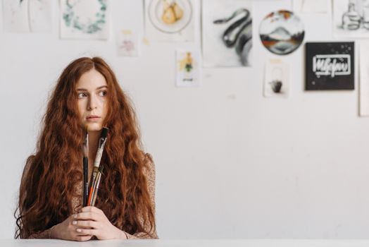 Young redheaded artist holding paintbrushes in art studio with artworks on the wall.