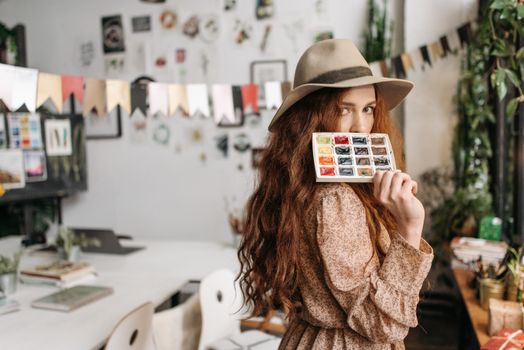 A woman artist in a hat holding a watercolor palette in a colorful studio environment.