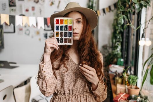 Artistic woman in a cozy studio holding a watercolor palette, surrounded by plants and creative decor.
