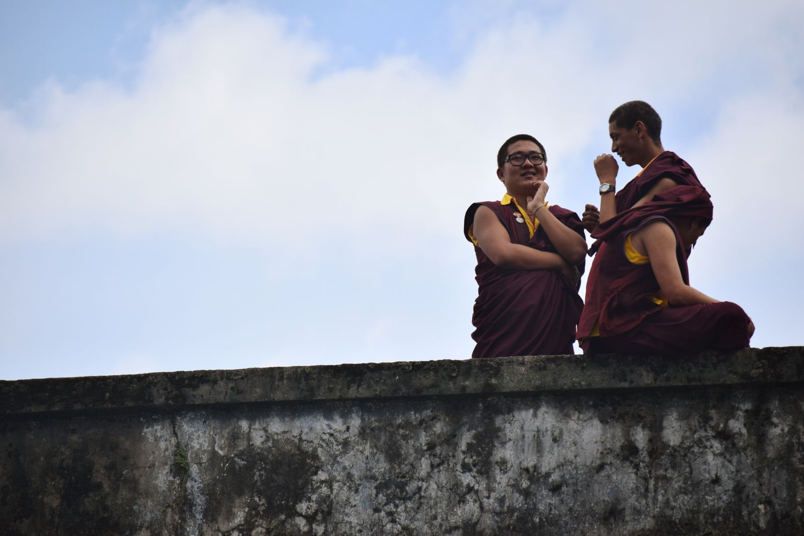 Two young monks in maroon robes laughing together on a monastery wall in Kalimpong, a pale cloudy sky behind them