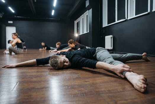 A group of adults practicing stretching exercises in a modern dance studio with wooden floors.