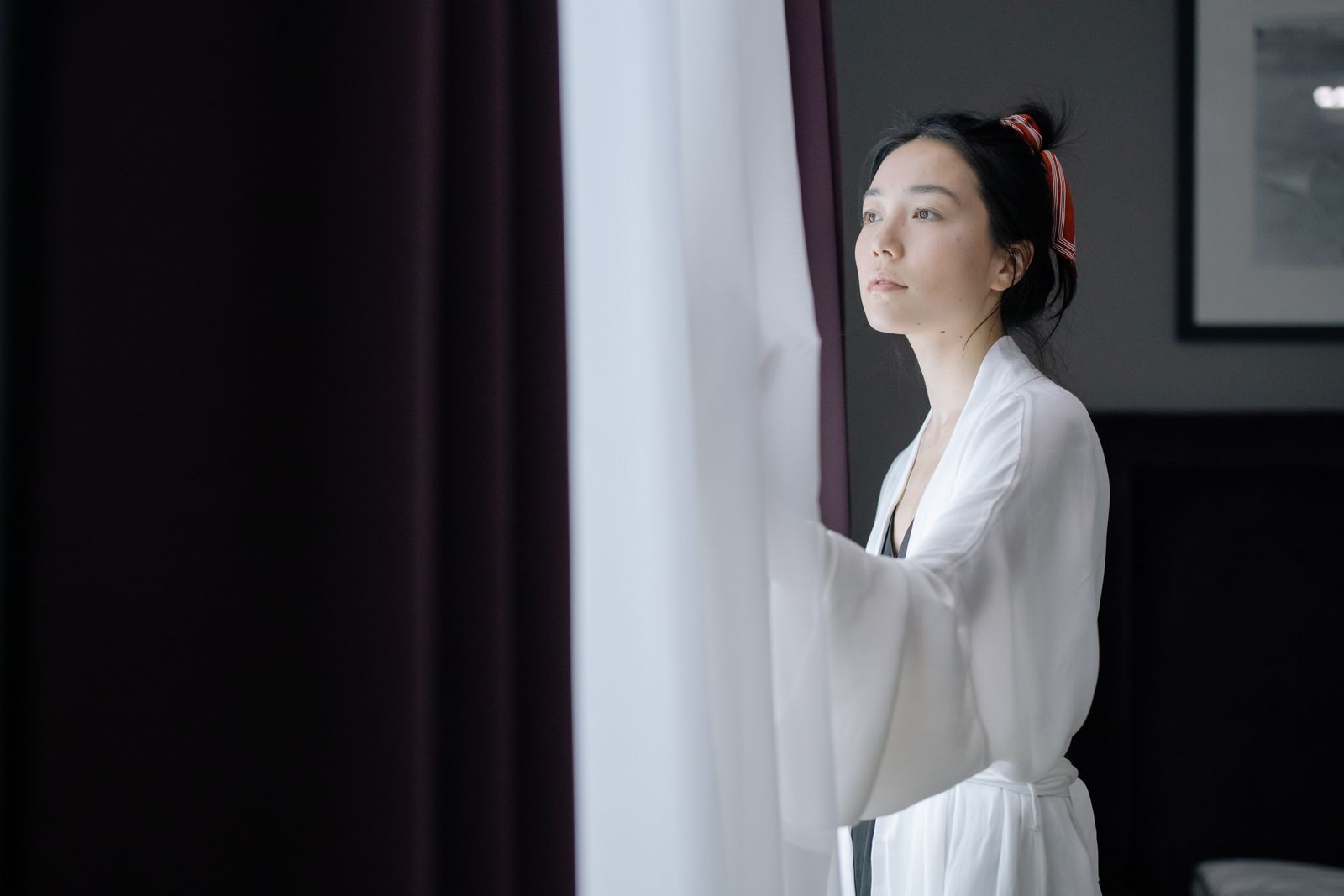 Woman in a white robe with a red hair tie gazing out a window beside sheer curtains