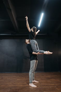 A couple performing a graceful yoga pose indoors with a wooden floor and dark backdrop.