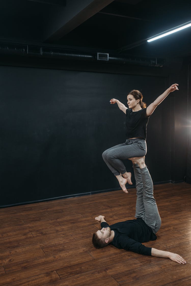 A Man Lying On A Wooden Floor While Lifting His Partner Using His Feet