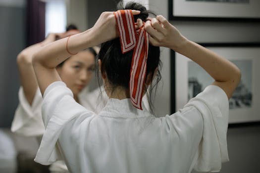 Close-up of a woman in a robe tying her hair with a red ribbon in front of a mirror indoors.