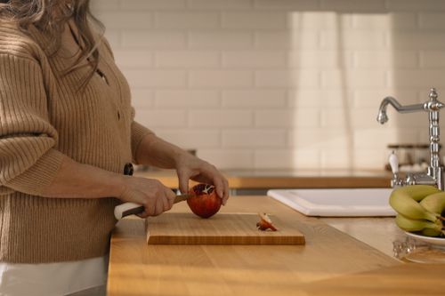 Free Senior woman slicing a pomegranate on a wooden cutting board in a sunlit kitchen setting. Stock Photo