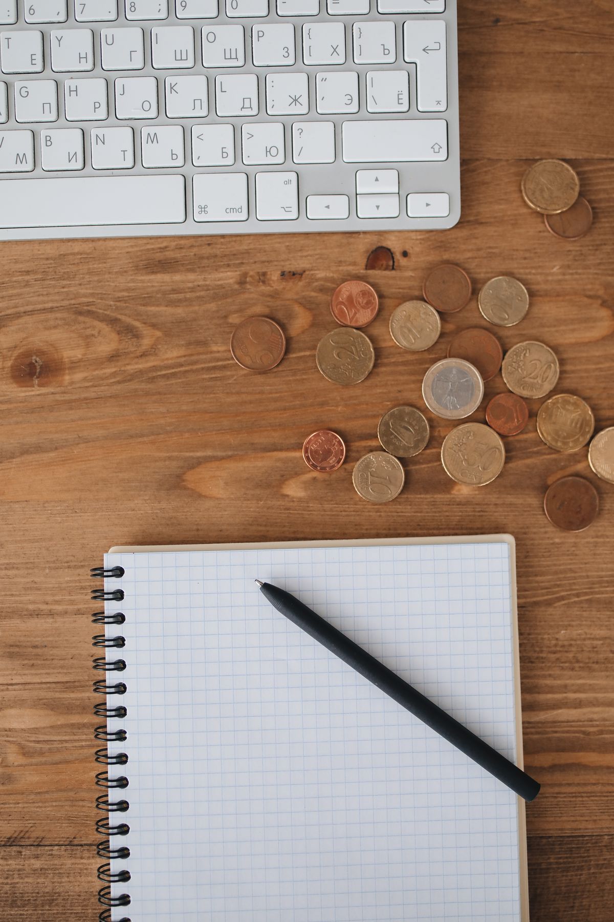 Vista de cima de mesa de trabalho com teclado, moedas e caderno de anotações, representando organização financeira pessoal