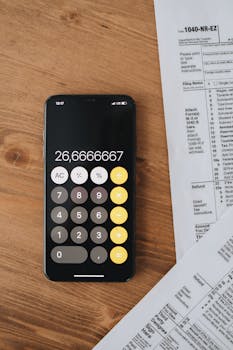 Overhead shot of a smartphone calculator displaying a number next to tax forms on a wooden surface.