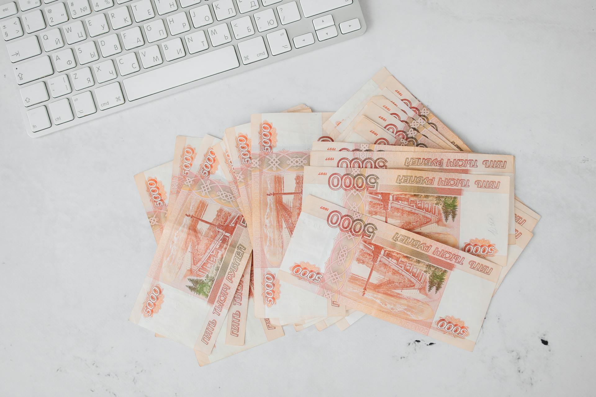 Pile of 5000 ruble banknotes next to a keyboard on a white surface, viewed from above.