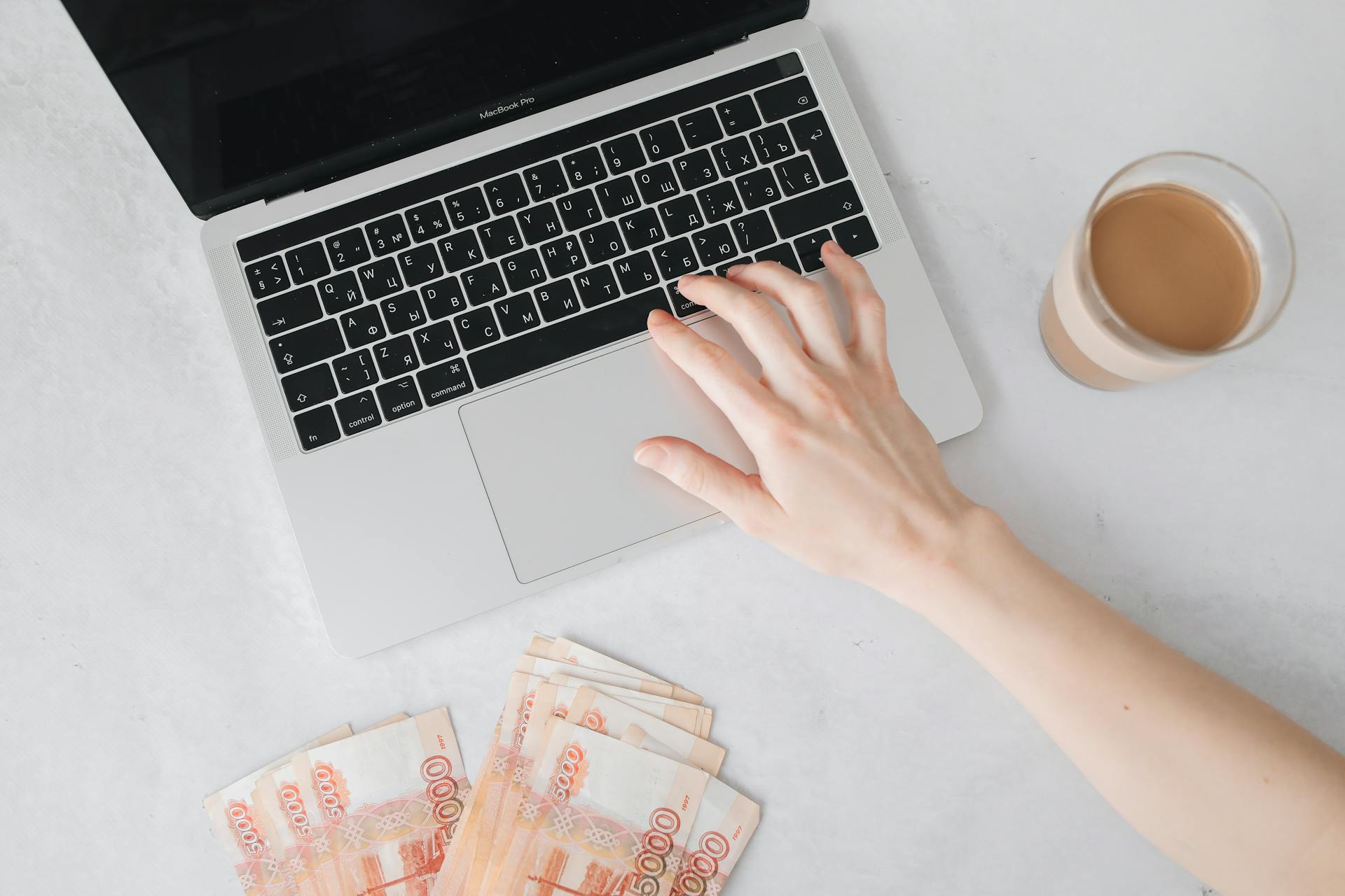 Close-up of a hand on laptop keyboard with cash and coffee on table.