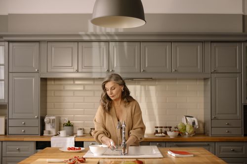 Free Senior woman at kitchen sink preparing ingredients amidst a modern setting, for cooking. Stock Photo
