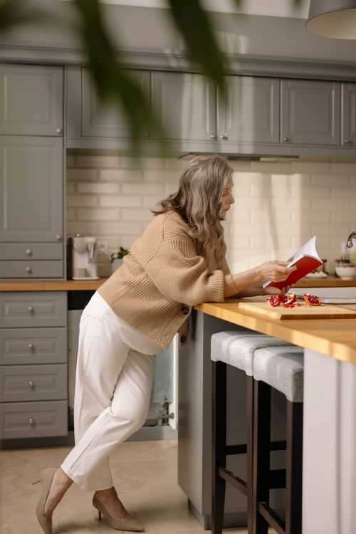 Free Elderly woman in kitchen reading a book, surrounded by a cozy atmosphere and fresh pomegranates. Stock Photo