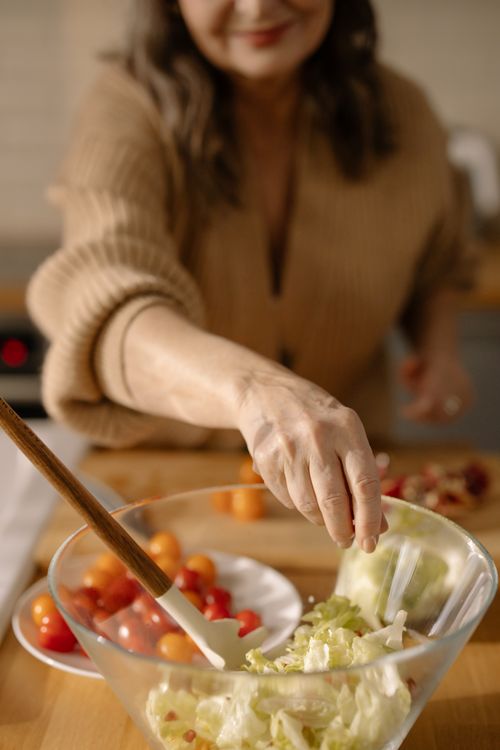 Free Woman preparing a fresh salad with tomatoes and lettuce in a glass bowl in a cozy kitchen setting. Stock Photo