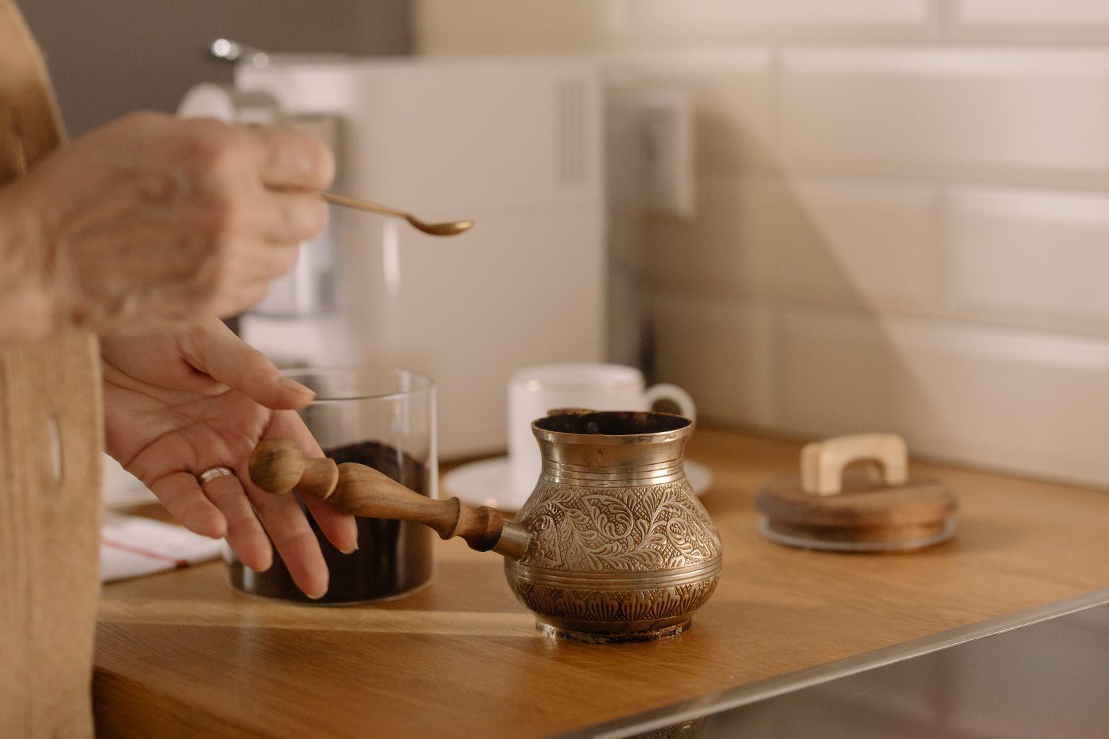 A hand lifting the lid of an engraved brass coffee pot beside a small glass