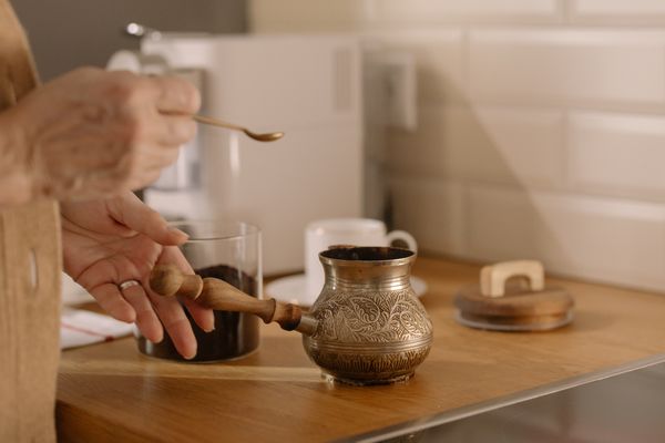 A hand lifting the lid of an engraved brass coffee pot beside a small glass