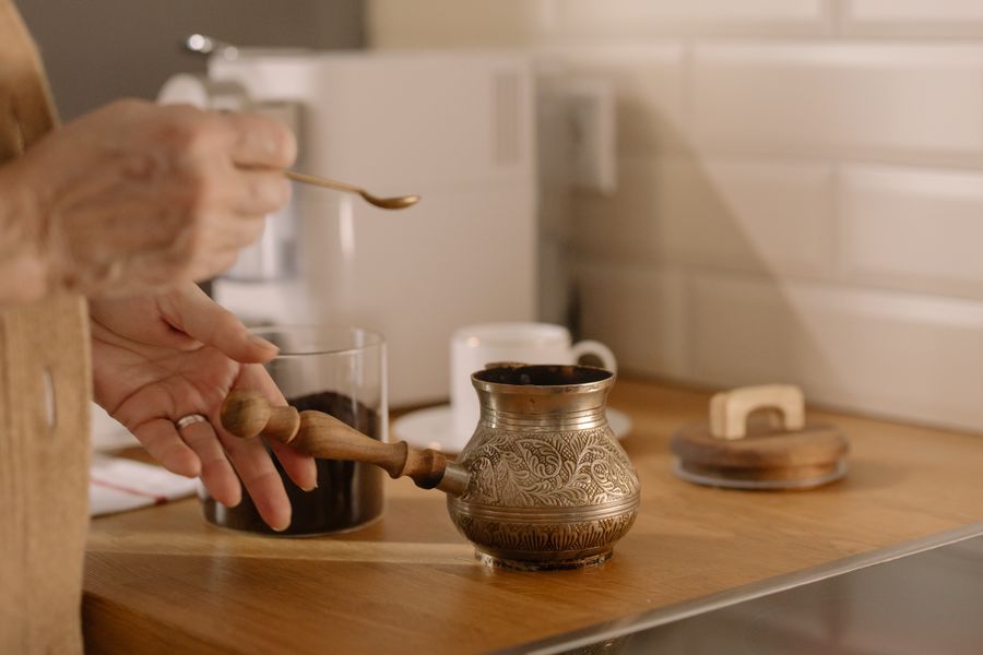A hand lifting the lid of an engraved brass coffee pot beside a small glass