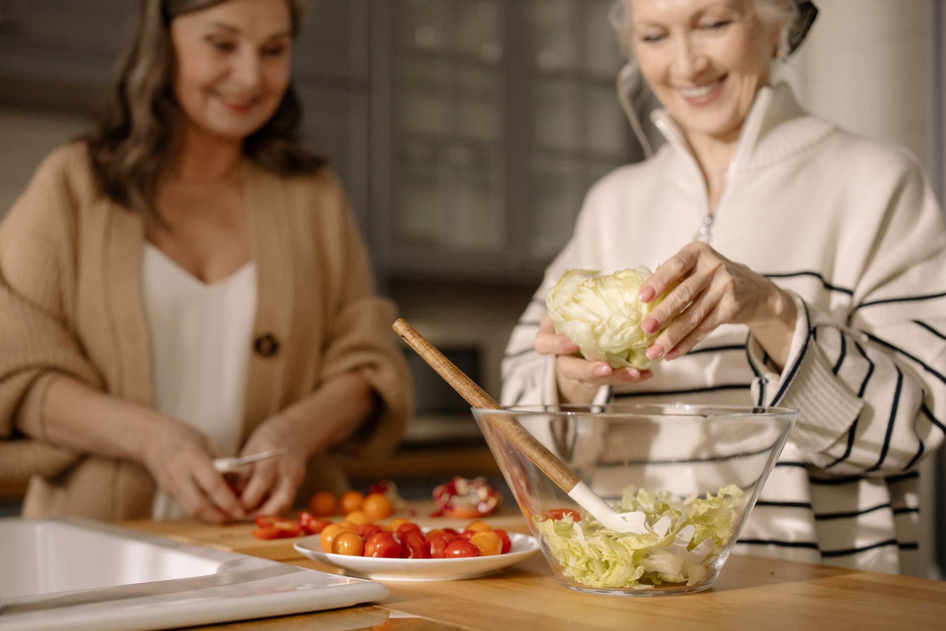 地中海飲食改善血糖:真實案例分享與實踐指南 Senior women happily preparing a fresh salad with cherry tomatoes and lettuce in a cozy kitchen setting.