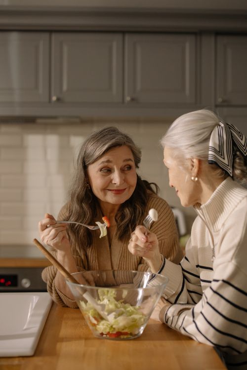 Free Two senior women share a light-hearted conversation over a fresh salad in a cozy kitchen. Stock Photo