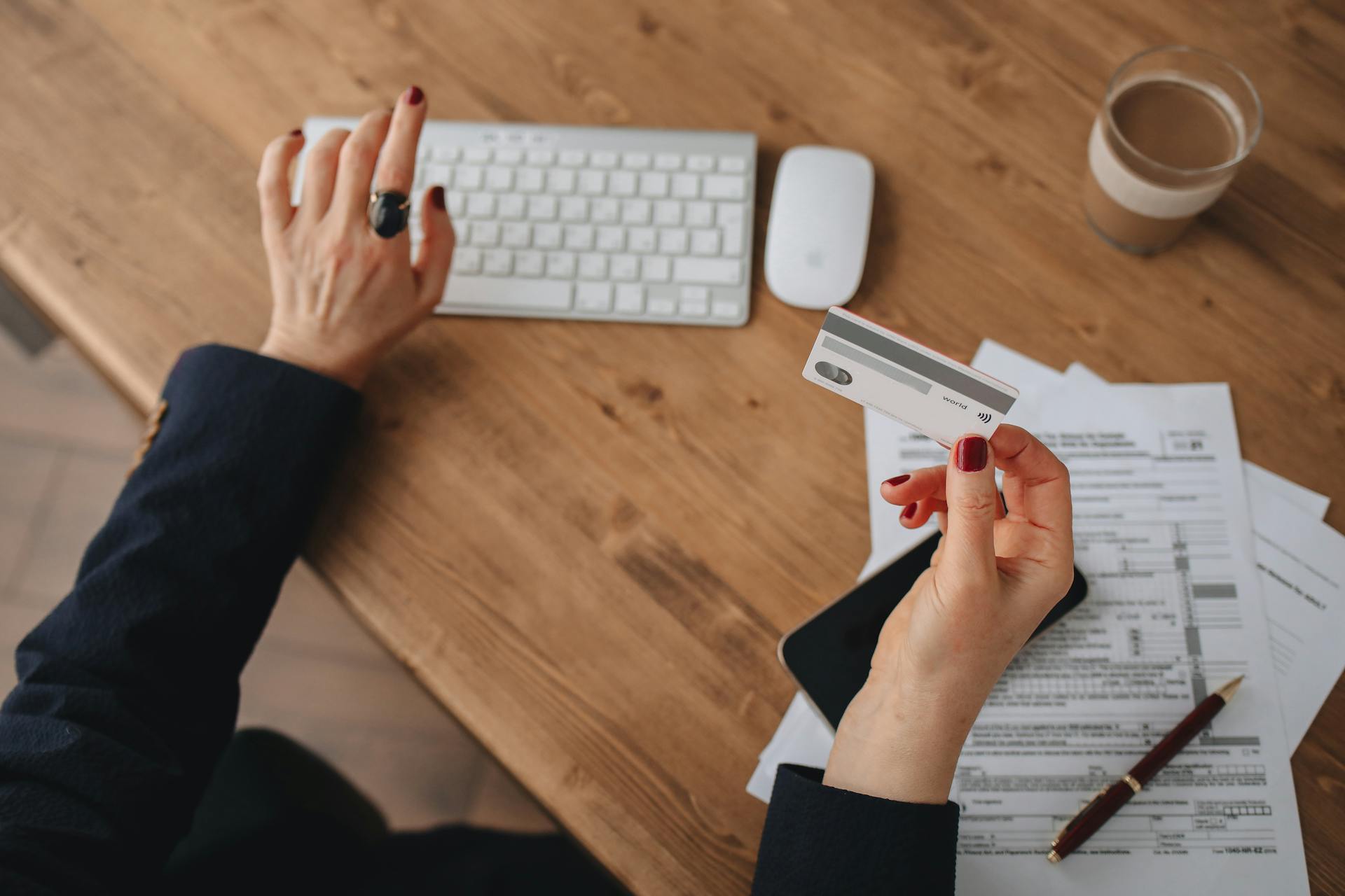 A woman uses an Apple Magic Keyboard while holding a credit card over paperwork on a wooden desk.