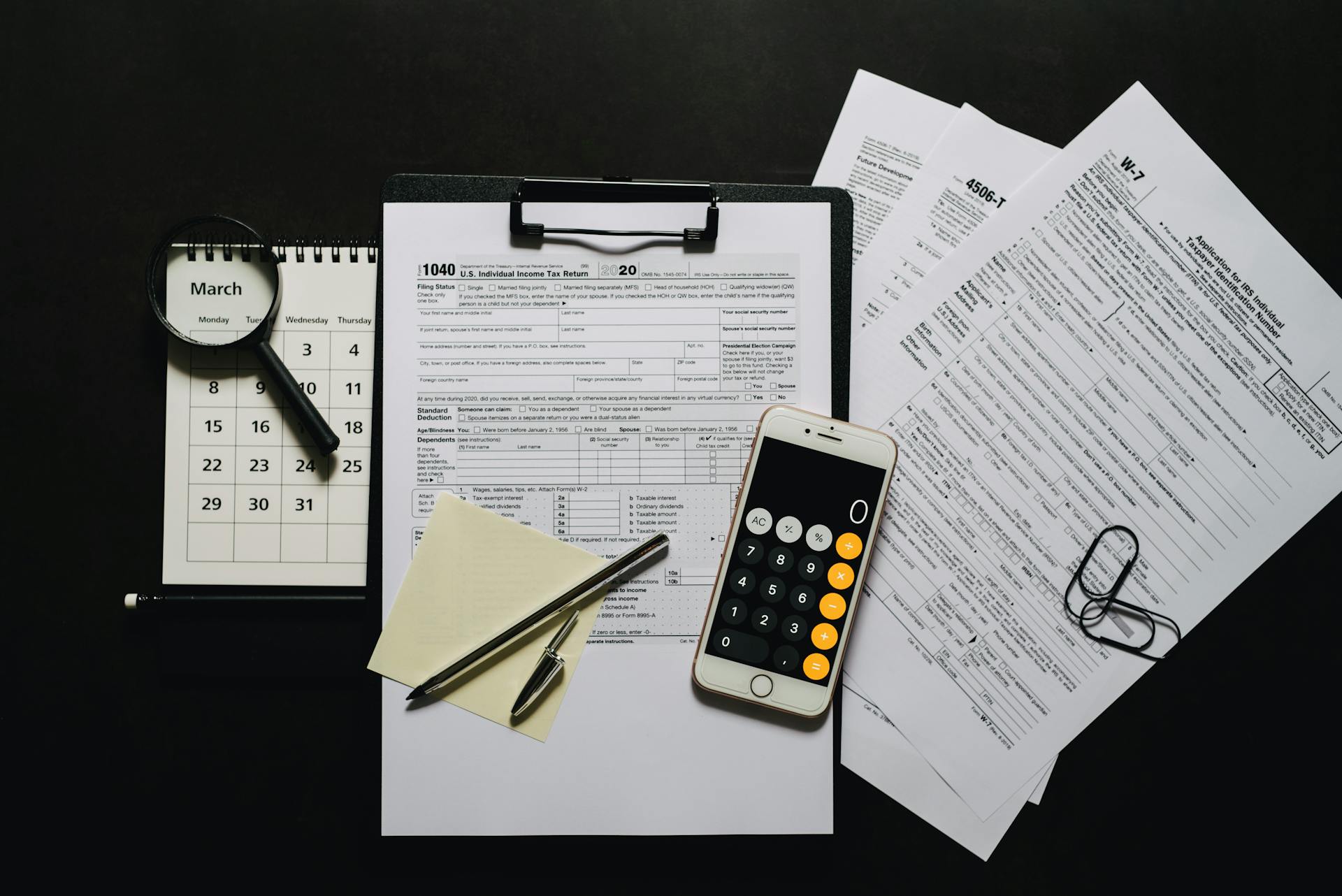 Top view of tax documents, calculator, magnifying glass, and calendar on a black surface.