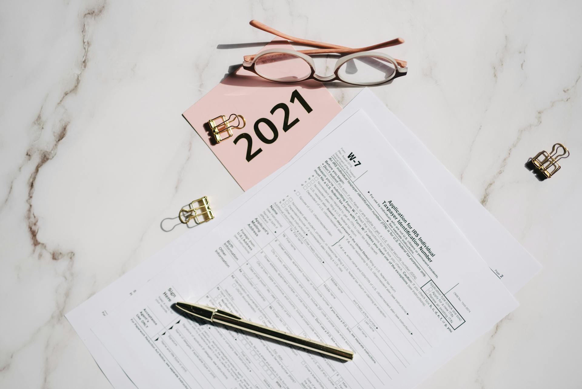 Close-up of W-7 tax forms with glasses and pen on a marble desk, ideal for finance concepts.