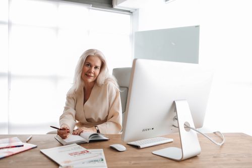 Free Elegant senior woman writing at office desk with computer, showcasing a professional work environment. Stock Photo