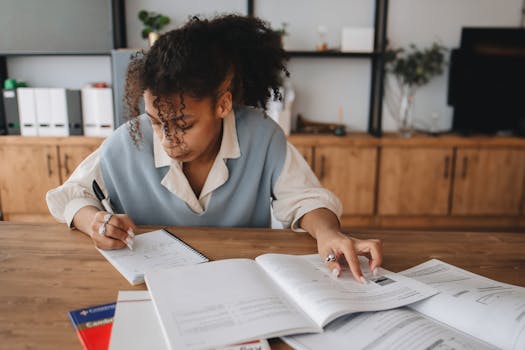 Focused young woman taking notes from a book at home, surrounded by study materials.