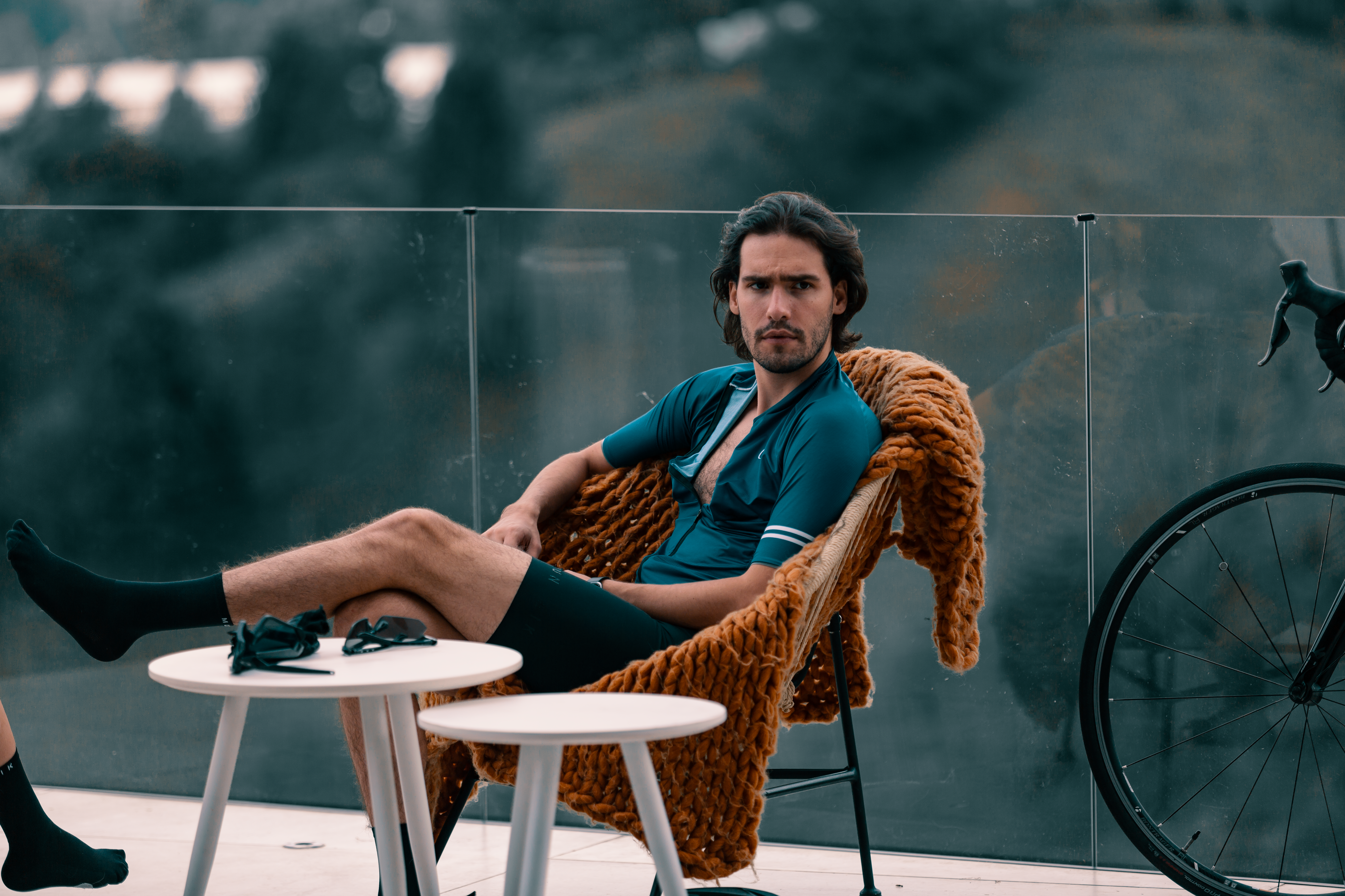 Man in cycling gear relaxing outdoors on a glass-railed terrace with a bicycle nearby.