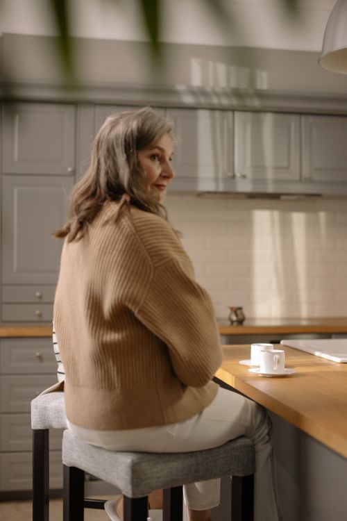 Free Senior woman sitting on a bar stool in a modern kitchen, enjoying a cup of coffee. Stock Photo