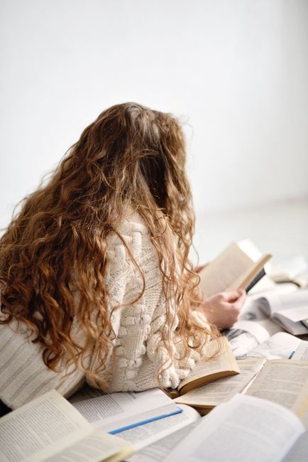 Back view of a woman with wavy hair reading amidst piles of open books.