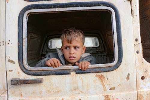 Scared ethnic child peeping out of aged transport hole with rust while looking away in daylight
