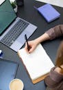 Hand of a Person Writing on a Notebook Near a Laptop