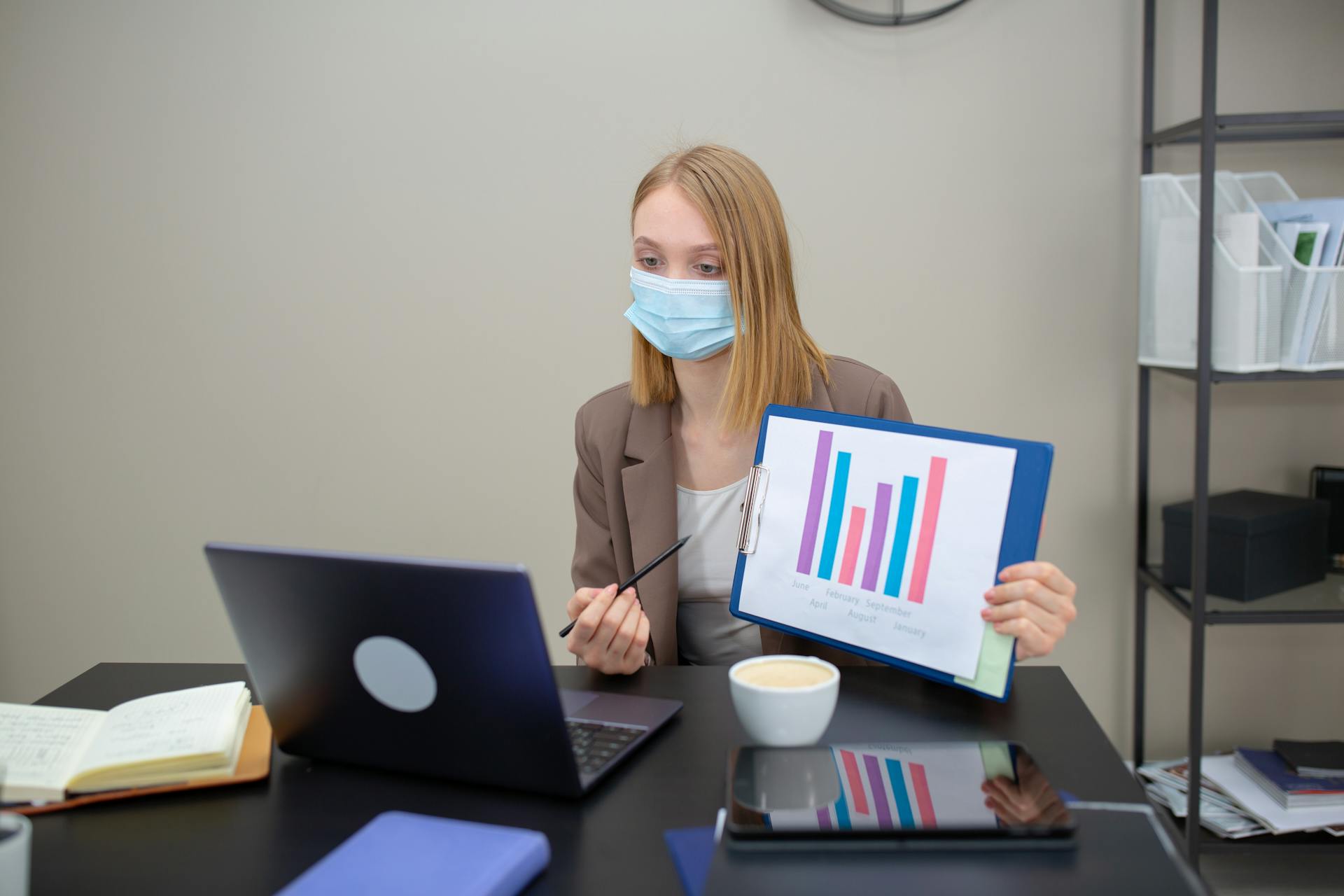 A woman in an office conducts a video call while presenting data with a mask on.