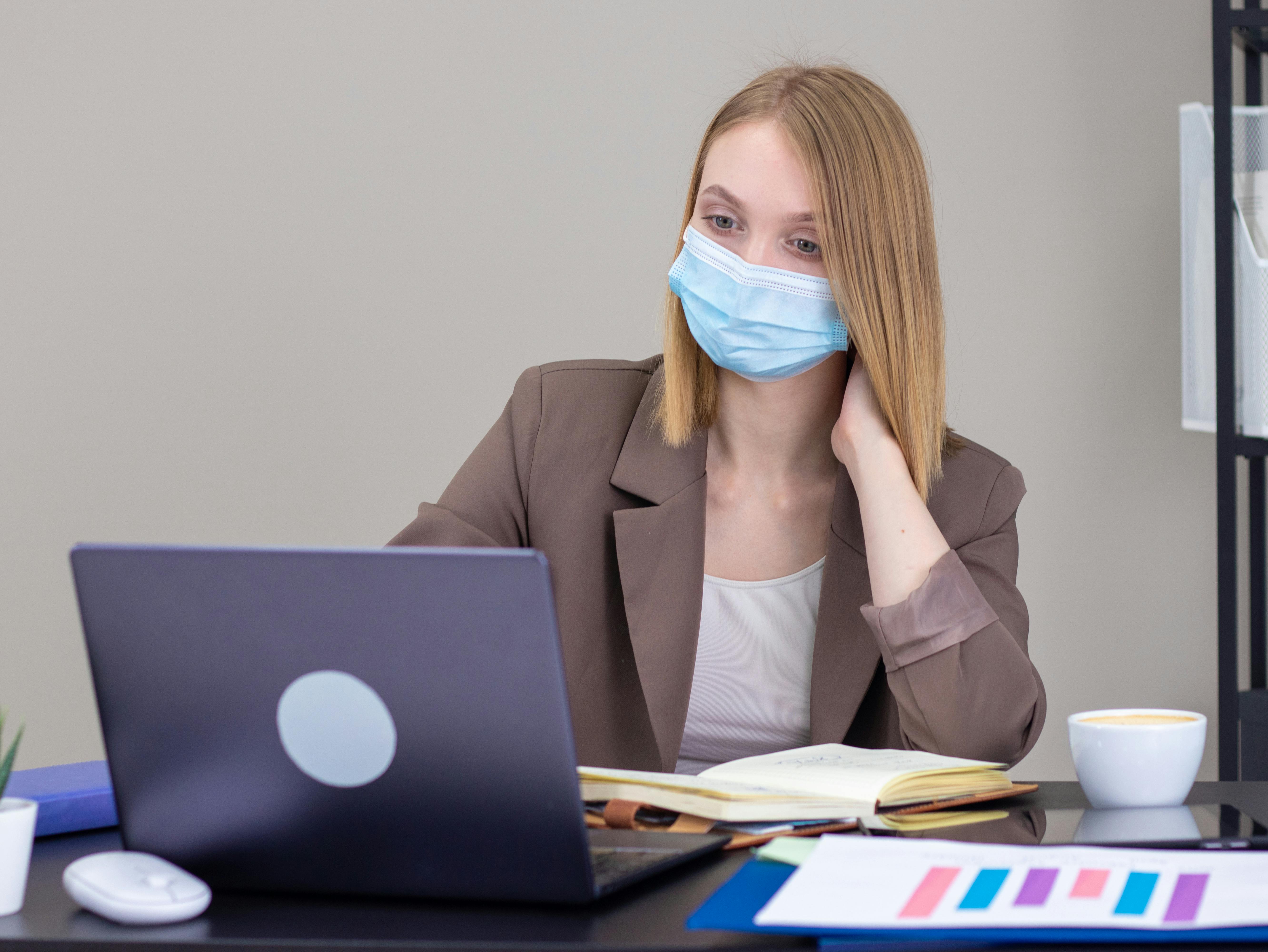Young caucasian woman in brown blazer wearing a face mask working on a laptop in an office environment.