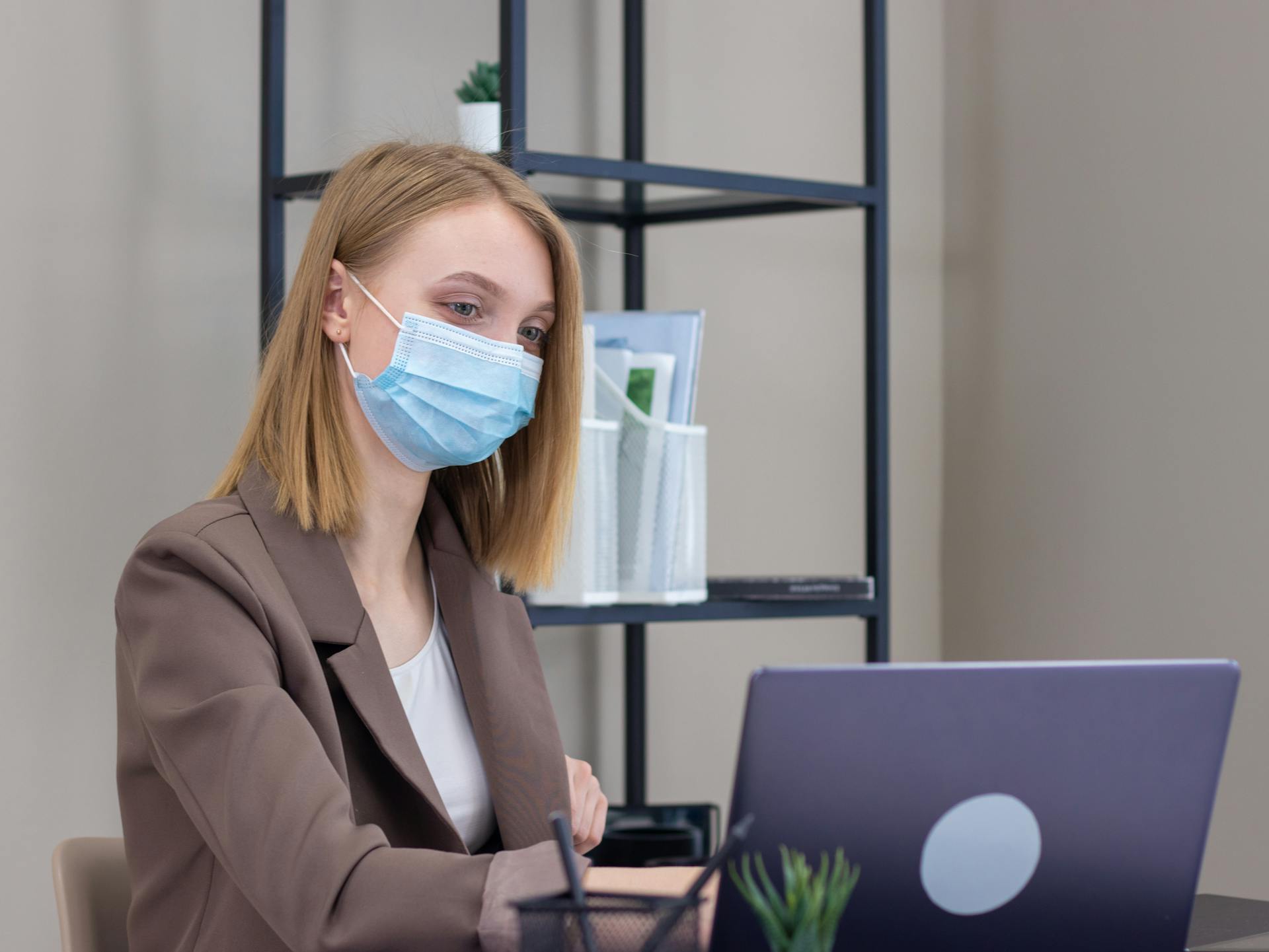 Professional woman working at office desk wearing face mask and using laptop for online tasks.